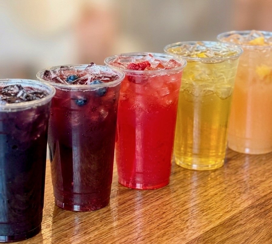 A lineup of five exotic lemonades — pomegranate, blueberry, raspberry, mango, and pineapple — in clear cups, colors ranging from deep purple to bright yellow